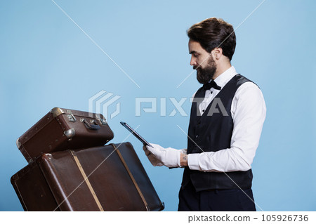 Hotel concierge posing with gadget and looking at online bookings to provide luxury assistance and services. Classy porter with gloves and tablet posing next to luggage, tourism assistance. Hotel concierge posing with gadget and looking at online bookings to provide luxury assistance and services. Classy porter with gloves and tablet posing next to luggage, tourism assistance. 105926736
