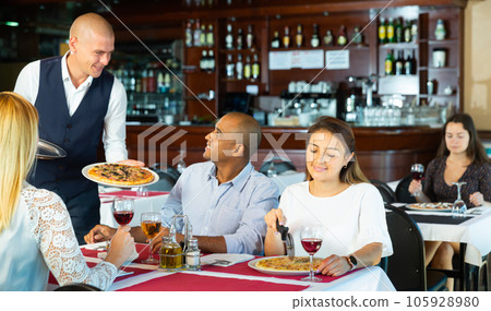 Polite waiter serving pizza to friendly company in pizzeria Polite waiter serving pizza to friendly company in pizzeria 105928980