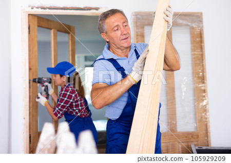 Middle-aged male carpenter in a blue uniform checking wooden planks indoors Middle-aged male carpenter in a blue uniform checking wooden planks indoors 105929209