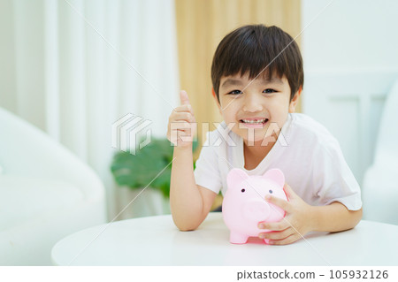 Happy cheerful Asian little young boy try to putting a coins into saving piggy and smiles to camera. Money saving learning in children concept. 105932126