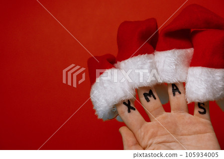 Fingers dressed in Santa-Claus red-white hats and word text XMAS on red background. Concept of Christmas or New Year holidays 105934005