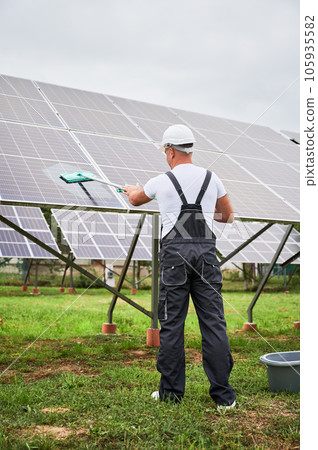 Professional worker cleaning solar PV panel. Man making sure solar batteries in good condition. Back view of worker in overalls and helmet cleaning panel by mop. 105935582