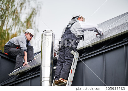 Installers installing photovoltaic solar panels on roof of house. Men engineers in helmets building solar module system with help of hex key. Concept of alternative, renewable energy. Installers installing photovoltaic solar panels on roof of house. Men engineers in helmets building solar module system with help of hex key. Concept of alternative, renewable energy. 105935583