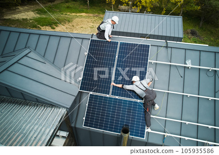 Builders installing photovoltaic solar panels on roof of house. Men engineers in helmets building solar module system with help of hex key. Concept of alternative, renewable energy. Aerial view. 105935586