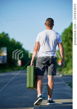 Young man walking along highway to nearest gas station to fill canister. Male adult needs to refuel stalled car. Back view of man walking near road with canister in hand. 105935655