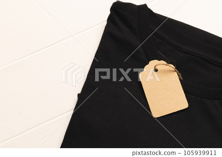 Close up of flat lay of black t shirt with tag and copy space on white boards background Close up of flat lay of black t shirt with tag and copy space on white boards background 105939911
