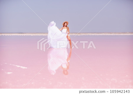 Woman in pink salt lake. She in a white dress and hat enjoys the scenic view of a pink salt lake as she walks along the white, salty shore, creating a lasting memory. Woman in pink salt lake. She in a white dress and hat enjoys the scenic view of a pink salt lake as she walks along the white, salty shore, creating a lasting memory. 105942143