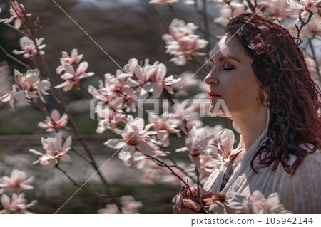 Magnolia park woman. Stylish woman in a hat stands near the magnolia bush in the park. Dressed in white corset pants and posing for the camera. 105942144