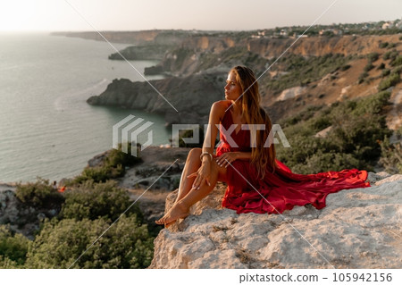 Woman sunset sea red dress, side view a happy beautiful sensual woman in a red long dress posing on a rock high above the sea on sunset. 105942156