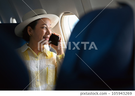 Positive middle aged woman tourist passenger talking on mobile phone and looking through airplane window. Positive middle aged woman tourist passenger talking on mobile phone and looking through airplane window. 105943269