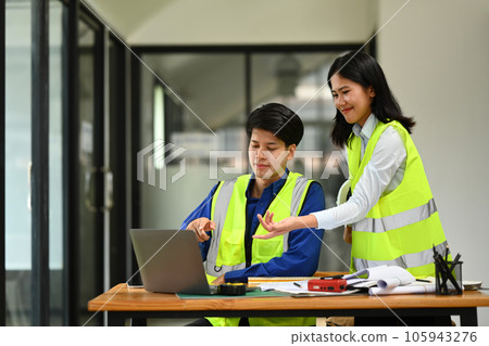 Two engineers in reflective vests working on blueprint and discussing the new project in office. 105943276