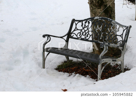 Black medal bench next to a tree in the snow Black medal bench next to a tree in the snow 105945882