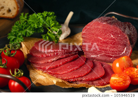 Whole and sliced bresaola on a metal round tray with tomatoes, garlic and parsley 105946519
