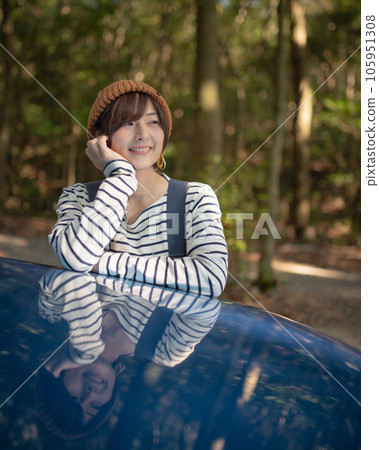 A woman looking into the distance with her chin resting on the hood of a blue car in the woods A woman looking into the distance with her chin resting on the hood of a blue car in the woods 105951308