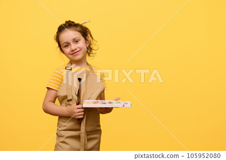Adorable baby girl in beige apron, talented painter posing with paintbrush and watercolors smiling looking at camera Adorable baby girl in beige apron, talented painter posing with paintbrush and watercolors smiling looking at camera 105952080