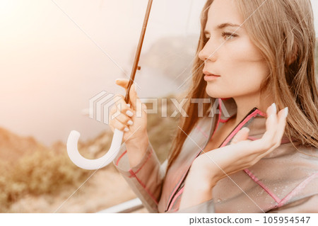 Woman rain umbrella. Happy woman portrait wearing a raincoat with transparent umbrella outdoors on rainy day in park near sea. Girl on the nature on rainy overcast day. 105954547