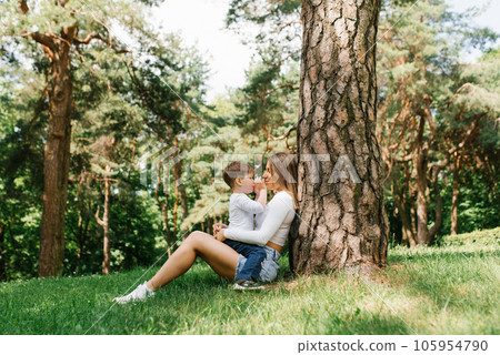 Young Caucasian mom and her three-year-old son have fun sitting in the park near a tree on the grass in summer Young Caucasian mom and her three-year-old son have fun sitting in the park near a tree on the grass in summer 105954790