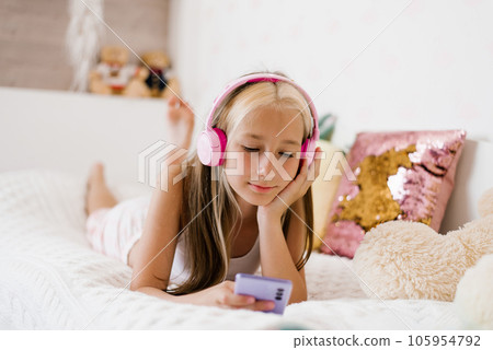Beautiful teenage girl is relaxing in bed using a touchscreen smartphone, listening to music with a wireless headset, surfing the Internet in the bedroom, relaxing comfortably, smiling, enjoying a Beautiful teenage girl is relaxing in bed using a touchscreen smartphone, listening to music with a wireless headset, surfing the Internet in the bedroom, relaxing comfortably, smiling, enjoying a 105954792