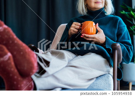 Cropped mature middle aged woman in sweater and warm socks relaxing in armchair with pumpkin shaped cup of hot coffee or tea drink. Cozy calm autumn holidays at home. Fall hygge mood concept. 105954938