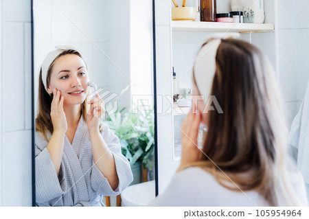 Young woman in bathrobe looking in mirror and making face massage with micro needle meso derma Roller. concept of natural procedures for skin care. Home Beauty self-care treatment. Selective focus. 105954964