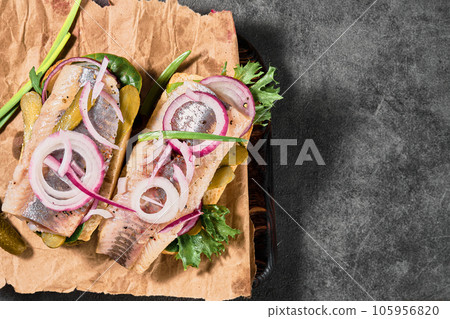 Dutch sandwich with herring, pickled cucumbers and red onions on a paper lining. Ingredients on a cutting board nearby. Layout on the table, selective focus and copy space Dutch sandwich with herring, pickled cucumbers and red onions on a paper lining. Ingredients on a cutting board nearby. Layout on the table, selective focus and copy space 105956820