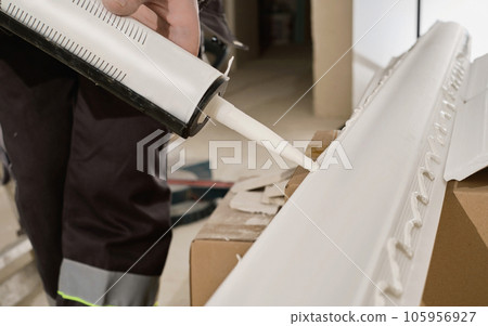 Master applies glue to the baseboard with a glue gun, hands close-up. The process of installing a ceiling plinth. 105956927