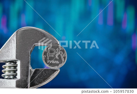 A coin with the symbol of the Russian ruble on a blurred blue background with a graph of falling quotes is squeezed with a wrench. News about the default and the collapse of the economy. Free text 105957830
