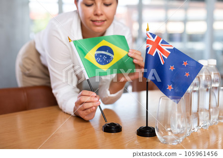 Young woman puts Brazil and New Zealand flags on table in office Young woman puts Brazil and New Zealand flags on table in office 105961456