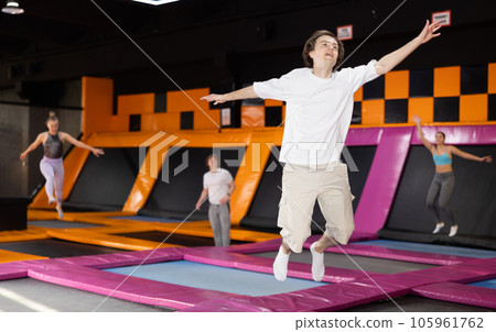 Smiling young male in white t-shirt and beige shorts having great time while jumping on colorful trampoline in game club 105961762