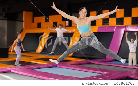 Excited young woman in blue top and gray leggings having great time while jumping on colorful trampoline in game club 105961763