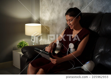 mother using tablet and breast pump machine to pumping milk for baby on sofa in the living room at night 105962933