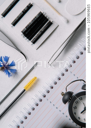 View from above of various tools for eyelash extension procedure, lying on table near cornflower and diary with clocks. 105969685