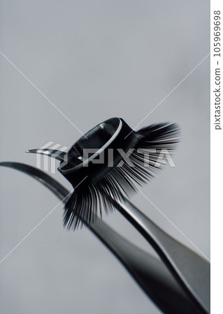 Close up of professional tweezers with ring of fluffy artificial eyelashes hanging on tip, against gray background. 105969698