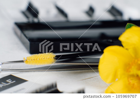 Flower slightly covering disposable eyelash extension brush lying on tweezers on table full of tools for procedure. Flower slightly covering disposable eyelash extension brush lying on tweezers on table full of tools for procedure. 105969707