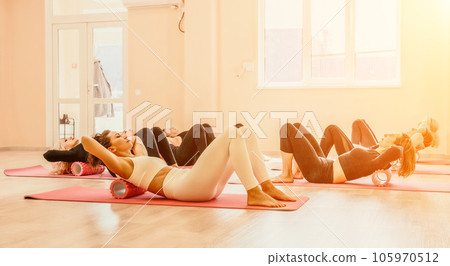Happy middle aged well looking women, performing fascia exercises on floor using massage foam roller - tool to relieve tension in the back and relieve muscle pain. Female fitness yoga routine concept Happy middle aged well looking women, performing fascia exercises on floor using massage foam roller - tool to relieve tension in the back and relieve muscle pain. Female fitness yoga routine concept 105970512