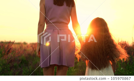 Mother and daughter walk holding hands across meadow grass at sunset backside view. Cute little daughter joins hands with young mother walking across field. Mother and daughter in field at holiday 105970867