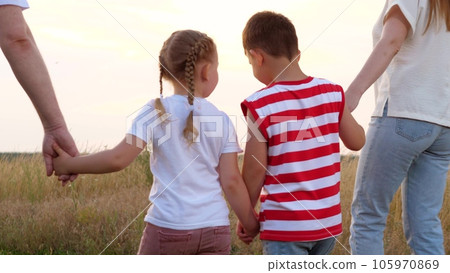 Happy little children walk joining hands with parents across summer meadow at sunset backside view. Loving children son and daughter discover nature with parents. Family and children walk in field Happy little children walk joining hands with parents across summer meadow at sunset backside view. Loving children son and daughter discover nature with parents. Family and children walk in field 105970869