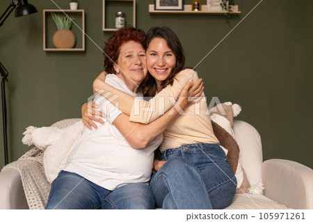 Portrait of a happy grandmother and granddaughter sitting on the sofa at home during the day, caring hugging and looking at the camera Portrait of a happy grandmother and granddaughter sitting on the sofa at home during the day, caring hugging and looking at the camera 105971261