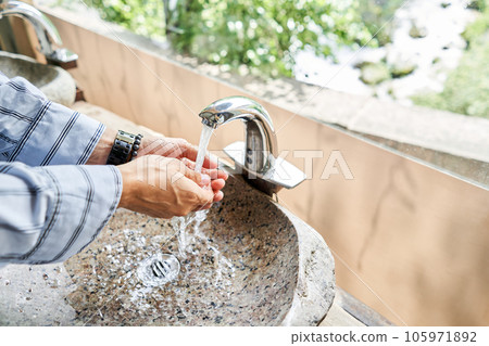 A man's hand touches the water in a beautiful sink with a metal faucet next to an mirror. Close-up of an elegant faucet in the bathroom sink next to stylish decorations. A man's hand touches the water in a beautiful sink with a metal faucet next to an mirror. Close-up of an elegant faucet in the bathroom sink next to stylish decorations. 105971892