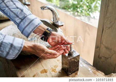 A man's hand touches the water in a beautiful sink with a metal faucet next to an mirror. Close-up of an elegant faucet in the bathroom sink next to stylish decorations. A man's hand touches the water in a beautiful sink with a metal faucet next to an mirror. Close-up of an elegant faucet in the bathroom sink next to stylish decorations. 105971893