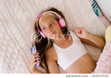 Beautiful teenage girl is relaxing in bed using a touchscreen smartphone, listening to music with a wireless headset, surfing the Internet in the bedroom, relaxing comfortably, smiling, enjoying a 105972430
