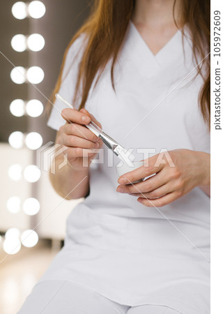 Female cosmetologist kneads a face mask in a bowl and holds a brush Female cosmetologist kneads a face mask in a bowl and holds a brush 105972609