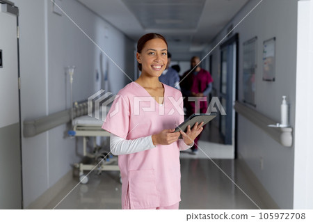 Portrait of happy biracial female doctor wearing scrubs holding tablet in corridor at hospital Portrait of happy biracial female doctor wearing scrubs holding tablet in corridor at hospital 105972708