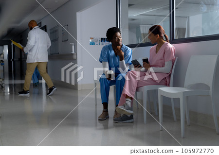 Diverse female doctors wearing scrubs drinking coffee and using smartphone in corridor at hospital Diverse female doctors wearing scrubs drinking coffee and using smartphone in corridor at hospital 105972770