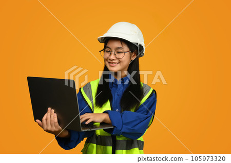 Smiling Asian girl wearing helmet holding laptop isolated on yellow background, future dream job career as engineer and architect 105973320