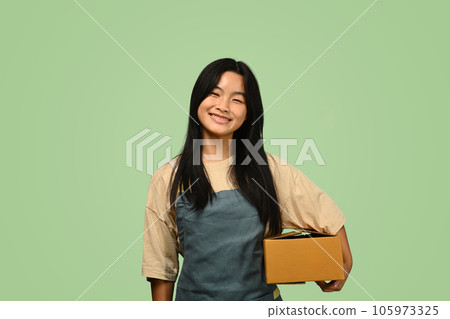 Cute teenage Asian girl holding parcel box isolated on green background. Post, package, shipping and delivery concept. Cute teenage Asian girl holding parcel box isolated on green background. Post, package, shipping and delivery concept. 105973325
