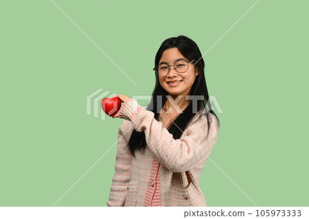 Smiling Asian teenage girl holding red heart isolated on green background. Love, valentine, gift and health symbol. 105973333