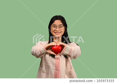 Smiling Asian teenage girl holding red heart isolated on green background. Love, valentine, gift and health symbol. Smiling Asian teenage girl holding red heart isolated on green background. Love, valentine, gift and health symbol. 105973335