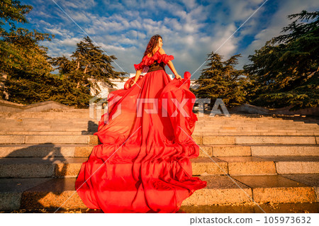 Sunrise red dress. A woman in a long red dress against the backdrop of sunrise, bright golden light of the sun's rays. The concept of femininity, harmony. 105973632