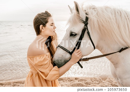 A woman in a dress stands next to a white horse on a beach, with the blue sky and sea in the background. A woman in a dress stands next to a white horse on a beach, with the blue sky and sea in the background. 105973650